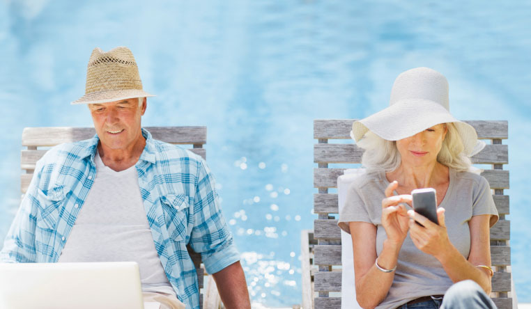 Senior couple sitting by swimmimg pool. Man looking at laptop. Woman looking at phone.